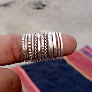 May include: A collection of silver rings with various designs, including textured, twisted, and floral patterns. The rings are stacked together, showcasing a variety of styles and textures. The rings are made of a shiny, metallic material.