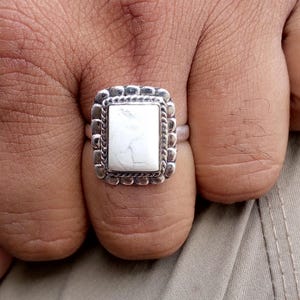 May include: A silver ring with a rectangular white stone. The stone is set in a decorative frame with a rope-like border. The ring has a textured, scalloped edge.