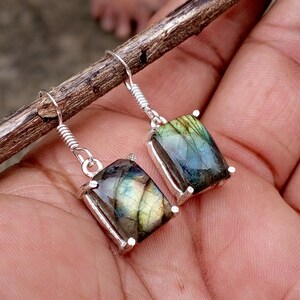 May include: A pair of silver drop earrings featuring rectangular labradorite gemstones. Each stone displays iridescent flashes of blue, green, and gold within a polished silver setting, suspended from simple silver ear wires with a coiled detail.