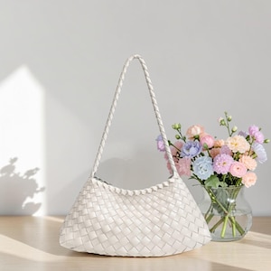 May include: A white woven shoulder bag with a braided handle. The bag has a curved shape and a textured surface. A vase of colorful flowers sits next to the bag on a wooden surface.