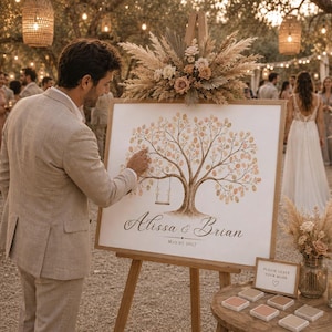 May include: A rectangular wedding guest book sign on a wooden easel featuring a painted tree with a swing, the names Alissa & Brian, and the date May 07 2027. The sign is decorated with dried pampas grass and flowers at the top.