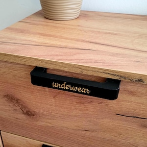 May include: Wooden dresser with black drawer pulls. The top drawer pull has the word "underwear" in gold lettering. A potted plant sits on top of the dresser.