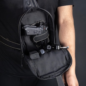 May include: Black sling bag with a concealed carry design. The bag is open, revealing a black handgun, a magazine, and other accessories. The bag has a zipper closure and a shoulder strap.