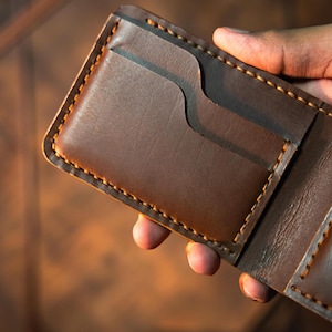May include: A brown leather wallet featuring a rectangular shape with rounded corners and visible thick tan stitching along the edges. The wallet includes a curved card slot opening on the front panel, showcasing a smooth, matte texture.