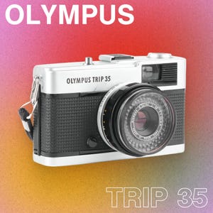 Op de afbeelding: Een Olympus Trip 35 camera met een zwart en zilverkleurig lichaam, zwarte lens en een zwarte riem. De camera heeft de tekst "OLYMPUS" bovenaan en "OLYMPUS TRIP 35" aan de voorkant. De tekst "TRIP 35" staat onderaan.