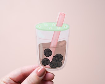 May include: A die-cut sticker of a clear glass filled with brown liquid and dark brown boba pearls shaped like dog faces. The glass has a light green lid decorated with small white daisies and a pink straw. The text little woman goods is visible.