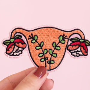 May include: Embroidered patch featuring a stylized uterus with floral details. The uterus is orange, the flowers are pink and red, and the leaves are green.