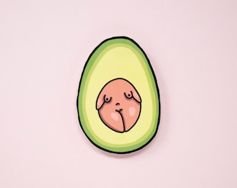 May include: A sticker illustration of a halved avocado with a light green flesh and a dark green outer skin. The central pit is replaced by a peach-colored, stylized human-like figure with a rounded torso, small breasts, and a navel.