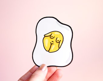 May include: A die-cut sticker shaped like a fried egg with a white oval-shaped egg white and a central yellow yolk. The yolk features a stylized illustration of a torso with two small dots representing breasts and a vertical line below.
