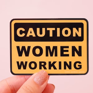 Könnte beinhalten: Ein gelbes Warnschild mit schwarzem Text, auf dem "ACHTUNG FRAUEN AM ARBEITSPLATZ" steht.