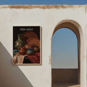 Puede incluir: Una impresión enmarcada que presenta una naturaleza muerta de cerámica y textiles, con el texto "FRIDA KAHLO" en la parte superior. La obra de arte incluye varios recipientes de cerámica marrones y verdes y telas estampadas. La impresión está montada en una pared blanca.