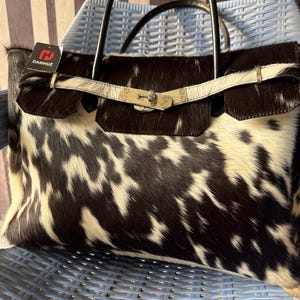 May include: A cowhide leather handbag with black and white spotted pattern. The bag has a flap closure with a silver-tone clasp and a white leather strap. Black handles arch over the top. A tag reads "DANHUZ".