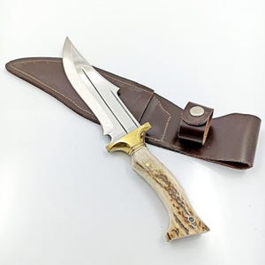 May include: A hunting knife with a stainless steel blade and a brass bolster. The handle is made of light brown antler material. The knife rests on a brown leather sheath with a belt loop.