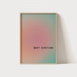 May include: A rectangular framed wall art piece featuring a soft, blurred gradient background that transitions from a central warm pink to orange and teal edges. The black text "DON'T OVERTHINK" is printed in a simple, handwritten font style.
