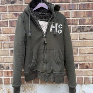May include: Olive green zip-up hoodie with a fleece-lined hood and interior. Features a front pocket and the letters "HS" on the chest. The jacket is hanging on a wooden hanger against a brick wall.