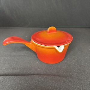 May include: A small, orange and red ceramic saucepan with a lid and a handle. The saucepan has a white spout and a rounded handle. The lid has a small, rounded handle. The color gradient goes from orange to red.