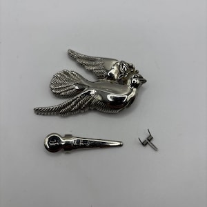 May include: A silver-toned dove brooch with outstretched wings, a separate pin, and a small spring. The brooch features intricate detailing on the bird's feathers and body. The pin has the text "L.A. Henry" engraved on it.
