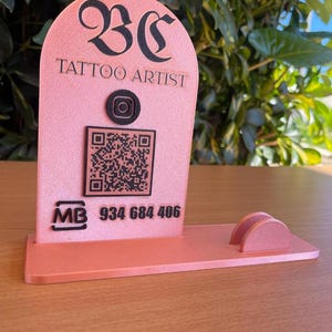 May include: A pink business card holder with the text "BC TATTOO ARTIST", a QR code, and a phone number. The holder has a slot for business cards and a decorative Instagram logo.