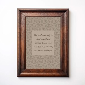 May include: A framed print with a dark brown wooden frame. The print has a beige background with a repeating pattern of leaves and flowers. A central beige rectangle contains the text: "The thief comes only to steal and kill and destroy; I have come that they may have life, and have it to the full."