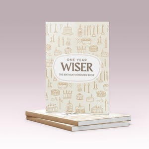 Può includere: Una pila di due libri con copertina beige chiaro. Il libro superiore presenta un motivo ripetuto di torte di compleanno e candele. Il testo "ONE YEAR WISER" è centrato sulla copertina, con "THE BIRTHDAY INTERVIEW BOOK" sotto.