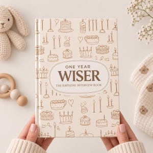 May include: A hardback book with a cream-coloured cover featuring brown line drawings of birthday cakes, candles, and hearts. The central oval contains the text "ONE YEAR WISER" and "THE BIRTHDAY INTERVIEW BOOK" in dark brown lettering.
