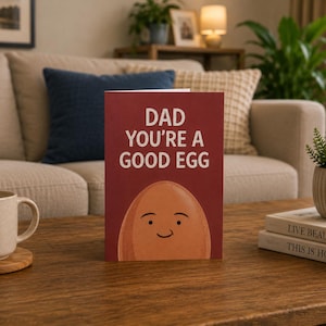 Puede incluir: Una tarjeta de felicitación granate con el texto "DAD YOU'RE A GOOD EGG" y un huevo de dibujos animados con una cara sonriente. Una taza beige y libros están sobre una mesa de madera. La escena está en un salón.