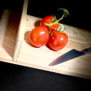 May include: Three ripe red tomatoes on a green vine rest on a light-colored wooden cutting board. A stainless steel knife is positioned near the tomatoes. The background is dark, creating a contrast with the bright red tomatoes.