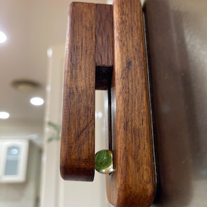 May include: A wooden refrigerator magnet with a dark brown finish. The magnet has a rectangular shape with a slot and a small, round, green marble embedded in the design. The magnet is attached to a stainless steel surface.