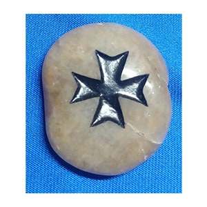 Puede incluir: Una piedra lisa, de forma ovalada, de color marrón claro. Una cruz de Malta negra está pintada en la superficie de la piedra. La piedra está sobre un fondo azul.