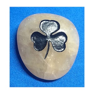 May include: A smooth, light-colored stone with a dark, metallic shamrock design. The stone is oval-shaped and sits against a blue background. The shamrock is a three-leaf clover, a symbol of luck and Irish heritage.