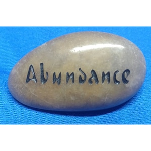 May include: A smooth, light brown stone with the word "Abundance" engraved in black lettering.