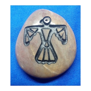 May include: A smooth, light brown stone with a black carved design of a stylised bird with outstretched wings.