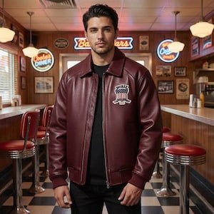 May include: Burgundy leather jacket with a ribbed hem and cuffs. The jacket features a front zipper closure and a patch on the chest with the text "AVIREX" and a patriotic design. The jacket is worn in a diner setting.