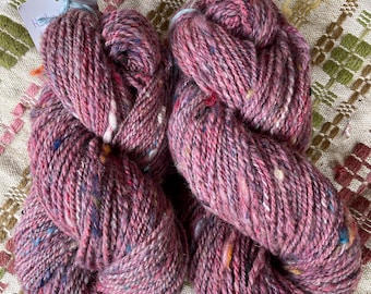 May include: Two skeins of pink yarn with flecks of blue, white, and orange. The yarn is thick and twisted, with a slightly textured appearance. The yarn is sitting on a patterned fabric with green, pink, and brown accents. A tag is attached to one skein.