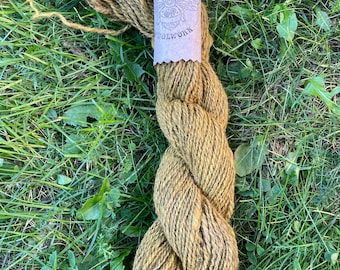 May include: A skein of olive green yarn rests on a bed of green grass. The yarn is twisted and has a paper label with the text "Full of Bees Woolwork" and a bee illustration. The yarn appears to be hand-dyed.