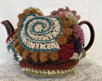 May include: A crocheted teapot cozy in shades of brown, green, cream, and burgundy. The cozy features a spiral design and various textures, including pom-poms and crocheted flowers.