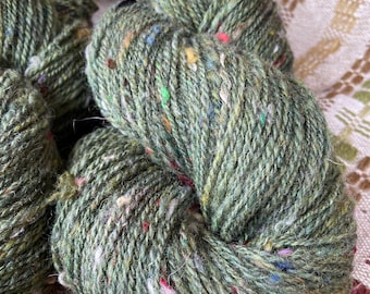 May include: Close-up of several skeins of green yarn with colorful flecks. The yarn appears to be a tweed or textured wool, with a slightly rough appearance. The yarn is arranged on a patterned fabric.