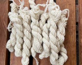 May include: Close-up of a hand-knitted off-white yarn swatch. The yarn is thick and twisted, creating a textured, braided appearance. The knitted piece is rectangular and lies on a wooden surface.