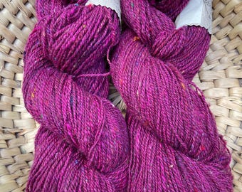 May include: Two skeins of vibrant magenta yarn with colorful flecks, presented in a woven basket. The yarn appears thick and textured, ideal for knitting or crochet projects. Each skein has a paper tag attached.