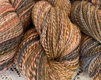 May include: Close-up of a skein of yarn in shades of brown, orange, and gray. The yarn is soft and fluffy, and it has a speckled appearance.