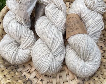 May include: A woven basket filled with several skeins of off-white yarn. Each skein is tightly wound and has a paper label with a bee illustration and the text "Full of Bees Woolwork". The basket is made of natural woven material.