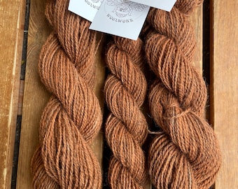 May include: Three skeins of brown wool yarn are displayed on a wooden surface. Each skein is twisted and has a tag that reads "Head Full of Bees Woolwork" with a bee illustration. The yarn has a natural, rustic appearance.