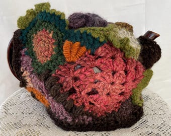 May include: A crocheted teapot cozy with a patchwork design in shades of green, brown, pink, and purple. The cozy is made of different crochet stitches and textures.