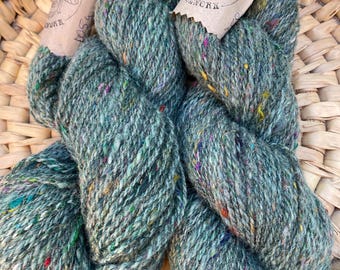 May include: Two skeins of teal-colored yarn with colorful flecks, labeled "Full of Bees" with a bee illustration. The yarn is thick and textured, perfect for knitting or crocheting projects. The yarn is displayed in a woven basket.