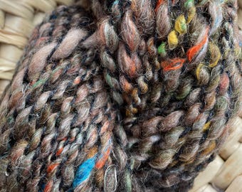 May include: Close-up of a ball of hand-spun yarn. The yarn is a mix of brown, tan, and black with colorful flecks of blue, orange, green, and yellow. The yarn has a thick, textured appearance, ideal for knitting or crocheting projects.
