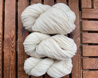 May include: Three skeins of off-white yarn are arranged on a wooden surface. The yarn is thick and appears soft, with visible strands. The wooden surface has a warm, brown tone, providing a contrasting backdrop for the yarn.