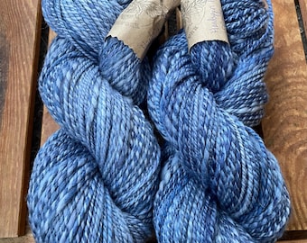May include: Two skeins of handspun yarn in varying shades of blue. The yarn is thick and textured, with a slightly mottled appearance. Each skein has a paper label wrapped around it. The yarn is displayed on a wooden surface.