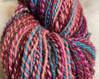 May include: A skein of yarn in shades of pink, purple, blue, and gray. The yarn is hand-spun and has a fuzzy texture.