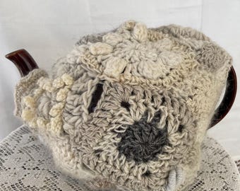 May include: A crocheted teapot cozy in beige and gray with a textured design. The cozy is made with different stitches and has a flower-like pattern on the top.
