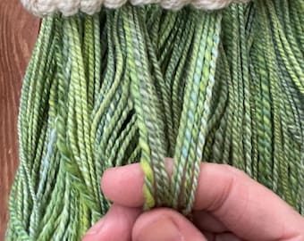 May include: Close-up of hand-dyed yarn in shades of green and yellow, with a cream-colored, twisted yarn accent. The yarn is held in a person's hand, showcasing its texture and color variations. Ideal for knitting and crochet projects.